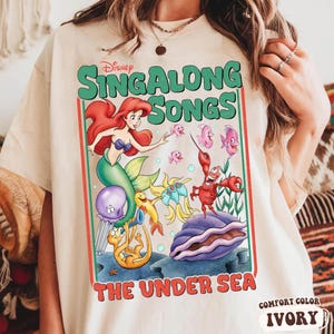 Pu&ograve; includere: T-shirt color avorio con una grafica colorata di "Sing Along Songs: The Under Sea" della Disney. Il design presenta Ariel, Flounder, Sebastian e altri personaggi. I testi "Sing Along Songs" e "The Under Sea" sono ben visibili.