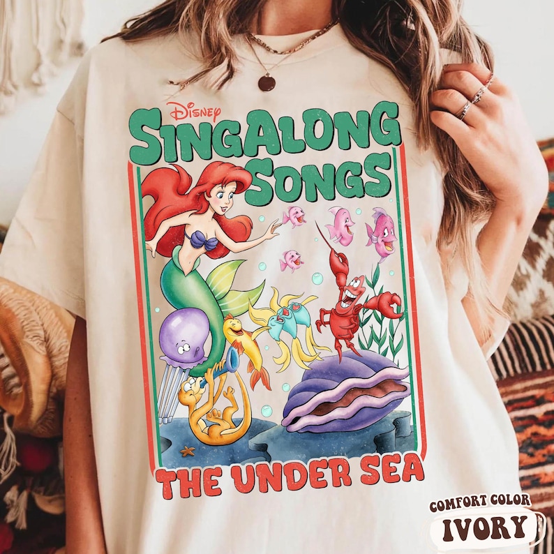 Pu&ograve; includere: T-shirt color avorio con una grafica colorata di "Sing Along Songs: The Under Sea" della Disney. Il design presenta Ariel, Flounder, Sebastian e altri personaggi. I testi "Sing Along Songs" e "The Under Sea" sono ben visibili.