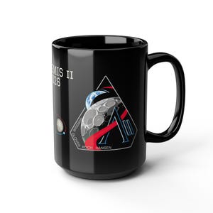 May include: Black ceramic mug featuring the Artemis II mission logo. The logo includes a stylized 'A' with a triangle, Earth, and moon graphic. The text "ARTEMIS II 26" is printed on the side, along with the names of the crew members.