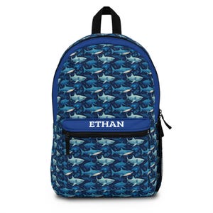 Peut inclure: Sac à dos bleu avec un motif de requins. Le sac à dos a un dessus et une poche avant bleus avec le nom "ETHAN" en blanc. Il comporte une poignée noire et une poche latérale.