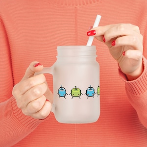Könnte beinhalten: Ein mattiertes Glas-Einmachglas mit Henkel, verziert mit einem pixeligen Design aus bunten Figuren. Ein weißer Strohhalm ist in das Glas gesteckt. Das Glas wird von einer Person gehalten, die einen korallenfarbenen Pullover trägt.