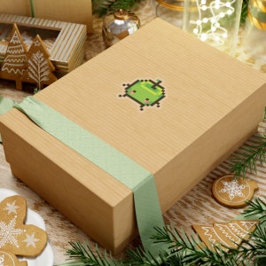 Könnte beinhalten: Eine hellbraune Geschenkbox mit einer grünen Pixelfigur auf der Vorderseite. Die Box ist mit einem hellgrünen Band gebunden. Die Box steht auf einem Tisch mit einer grünen Pflanze im Hintergrund.