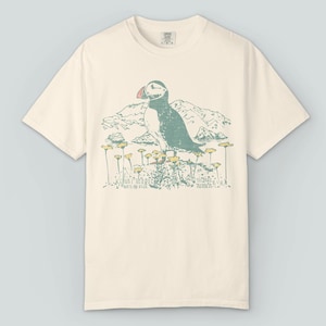 May include: Cream-colored t-shirt featuring a graphic of a puffin bird with a red beak, perched on a field of yellow flowers. The design includes mountains in the background and text that reads "Kenai Fjords, Alaska."