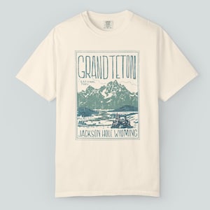 May include: Cream-colored t-shirt featuring a rectangular graphic of the Grand Teton National Park in Wyoming. The graphic includes the text "GRAND TETON" and "JACKSON HOLE WYOMING" with a mountain and lake scene in shades of blue and green.