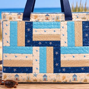 Peut inclure: Un sac fourre-tout matelassé avec un motif géométrique de bandes de tissu bleu clair, bleu foncé et beige. Le design présente des motifs imprimés de petites cabines de plage, de mouettes et d'étoiles de mer blanches. Deux anses en nylon bleu foncé.