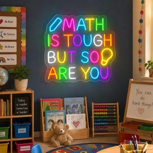 May include: A multi-colored LED neon sign mounted on a dark wall featuring the text "MATH IS TOUGH BUT SO ARE YOU" with a pencil icon at the top left and a lightbulb icon at the top right, all enclosed within a clear acrylic backing.