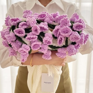 May include: A handmade crochet bouquet featuring lavender-colored bell-shaped flowers with green stems and leaves. The bouquet is held over a white knitted blanket with a tag that reads "Hand-made".