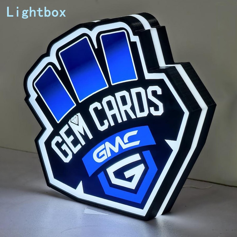 Pode incluir: Um letreiro luminoso LED de formato personalizado com um esquema de cores azul e preto. O design exibe o texto GEM CARDS com um &iacute;cone de diamante e a sigla GMC acima de uma letra G grande. O letreiro tem uma moldura preta espessa.