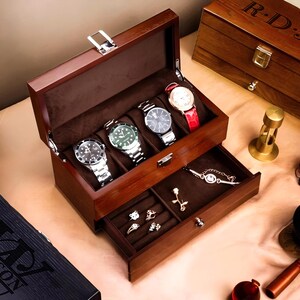 May include: A wooden watch box with a hinged lid and a pull-out drawer. The box has a brown finish and is lined with a dark brown velvet material. The top compartment has five slots for watches, and the bottom drawer has a ring holder and a small compartment for jewelry. The box is closed and the lid is slightly ajar.