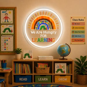 Puede incluir: Un letrero de neón LED circular que presenta un arcoíris colorido, una pequeña ilustración de una oruga verde y el texto "We Are Hungry FOR LEARNING" en letras multicolores, montado en una pared sobre una estantería de madera.