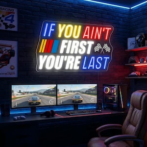 May include: A neon sign reading "IF YOU AIN'T FIRST YOU'RE LAST" is mounted on a brick wall. Two computer monitors below show a racing game. A gaming PC and a brown leather chair are also visible.