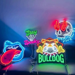 Pode incluir: Quatro letreiros de n&eacute;on: uma boca azul com l&iacute;ngua vermelha, um letreiro verde e rosa com a inscri&ccedil;&atilde;o Stay Spooky e uma m&atilde;o esquel&eacute;tica, um rosto de bulldog com a palavra BULLDOG em verde, e um cr&acirc;nio com chamas e o texto EVERYTHING'S FINE.