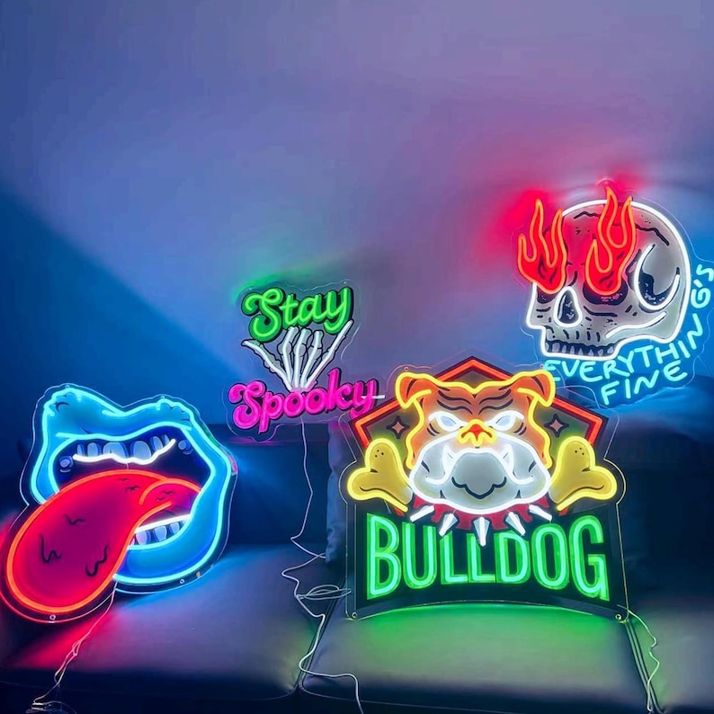 Pode incluir: Quatro letreiros de n&eacute;on: uma boca azul com l&iacute;ngua vermelha, um letreiro verde e rosa com a inscri&ccedil;&atilde;o Stay Spooky e uma m&atilde;o esquel&eacute;tica, um rosto de bulldog com a palavra BULLDOG em verde, e um cr&acirc;nio com chamas e o texto EVERYTHING'S FINE.