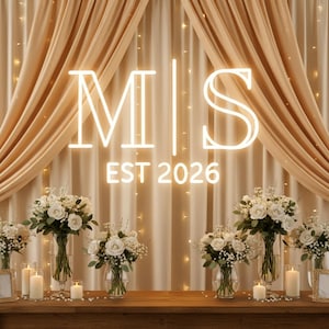 Puede incluir: Un telón de fondo para bodas o eventos con un letrero de neón brillante con las letras "MS" y el año "EST 2026". El letrero está colocado sobre un fondo de tela drapeada y luces de cadena. Arreglos florales blancos y velas adornan la mesa de madera.
