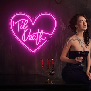 Peut inclure: Enseigne au néon rose vif en forme de cœur avec l'inscription "'Til Death" en cursive. L'enseigne est fixée sur un mur sombre. L'image présente également une femme en robe sombre.