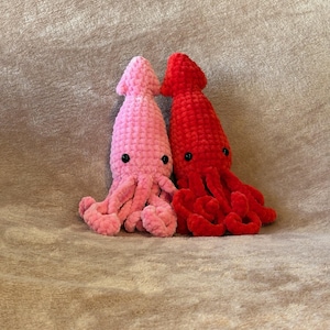 May include: Two handmade crochet squid plushies. One is light pink, the other is red. Both have black button eyes and curled tentacles. The plushies are made of a soft, textured yarn.
