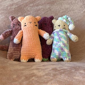 May include: Four crocheted cat plushies in different colors and patterns. The plushies are arranged in a row, with the orange cat in the center. The cat on the far right is wearing a blue and green striped sleep mask.