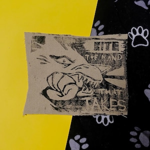 Peut inclure: Un patch de tissu carré beige avec une illustration noire d'un loup qui grogne et le texte "BITE THE HAND THAT TAKES". Le patch est posé sur un fond jaune et noir avec des empreintes de pattes.