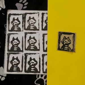 Könnte beinhalten: Eine Sammlung von Schwarz-Weiß-Blockdrucken mit einer stilisierten katzenartigen Figur. Die Drucke sind in einem Raster auf weißem Papier angeordnet, mit einem einzelnen Druck auf gelbem Hintergrund. Der Hintergrund enthält ein schwarz-weißes Pfotenmuster.