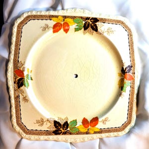 May include: Square, cream-coloured ceramic plate with a decorative border and colourful leaf accents. The plate has a brown border with a repeating pattern and a scalloped edge. The leaves are in shades of red, yellow, green, and black.