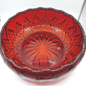 May include: A deep red glass bowl featuring a scalloped rim and intricate pressed patterns. The interior base displays a radiating starburst design, while the sides are decorated with a repeating diamond motif that catches the light.