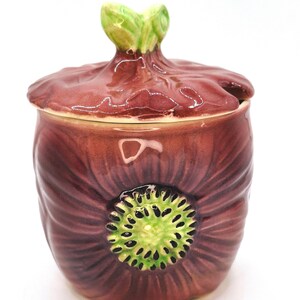 May include: A ceramic lidded jar shaped like a dark purple flower with a green centre and a green leaf-shaped handle on the lid. The jar features textured petals radiating from the centre and a glossy finish throughout the entire piece.