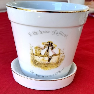 May include: White ceramic planter with a gold rim and a painted image of a girl carrying a basket. The planter has the text "To the house of a friend the way is never long..." printed on it.