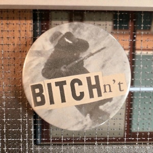 May include: A round button with a black and white image of a person holding a weapon. The button has the text "BITCH n't" in a bold, blocky font.