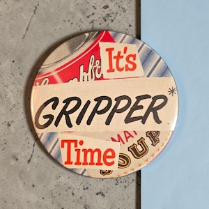 Puede incluir: Un botón redondo con las palabras "GRIPPER Time UP" en negro y rojo. El botón tiene un diseño de collage con elementos rojos y blancos, y un fondo a rayas azules y blancas.