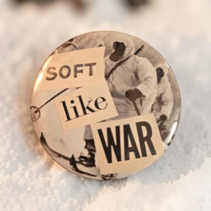 Peut inclure: Un bouton rond à l'esthétique vintage. Il présente une image en noir et blanc de personnes en tenue d'hiver, avec les mots "SOFT like WAR" superposés dans un style collage. Le bouton est posé sur un fond blanc et neigeux.