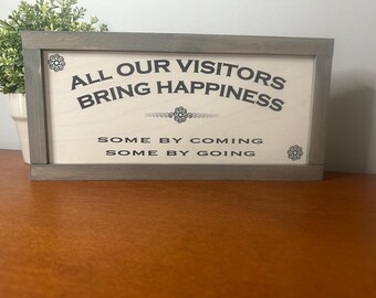 May include: A gray wooden sign with the text "All our visitors bring happiness, some by coming, some by going".