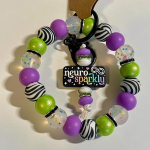 Puede incluir: Un llavero de cuentas con un cierre negro y un dije que dice "neuro sparkly". El llavero presenta una variedad de cuentas, incluyendo cuentas con estampado de cebra, verde lima, transparentes con motas coloridas y moradas s&oacute;lidas. Tambi&eacute;n hay un peque&ntilde;o dije de cupcake.