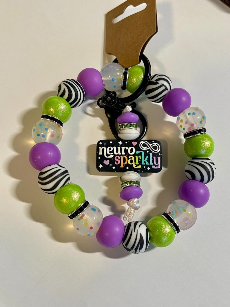 Puede incluir: Un llavero de cuentas con un cierre negro y un dije que dice "neuro sparkly". El llavero presenta una variedad de cuentas, incluyendo cuentas con estampado de cebra, verde lima, transparentes con motas coloridas y moradas s&oacute;lidas. Tambi&eacute;n hay un peque&ntilde;o dije de cupcake.