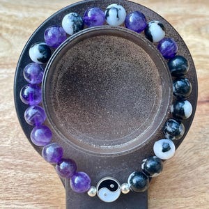 May include: A beaded bracelet featuring alternating purple and black and white speckled beads. The bracelet includes a silver charm with a yin-yang symbol. The bracelet is displayed on a dark gray stand.