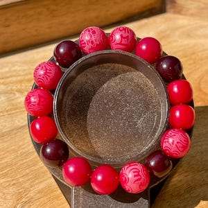 May include: A beaded bracelet featuring a mix of red and burgundy beads. Some beads have a carved design, while others are solid. The bracelet is displayed on a dark metal stand, showcasing its colors and textures.