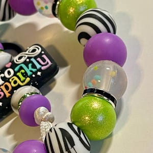 Puede incluir: Una pulsera de cuentas con una mezcla de cuentas redondas de varios colores y dise&ntilde;os. La pulsera incluye cuentas moradas, verdes, transparentes y con estampado de cebra en blanco y negro. Tambi&eacute;n es visible un dije con el texto "So Sparkly".