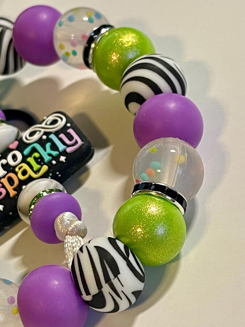 Puede incluir: Una pulsera de cuentas con una mezcla de cuentas redondas de varios colores y dise&ntilde;os. La pulsera incluye cuentas moradas, verdes, transparentes y con estampado de cebra en blanco y negro. Tambi&eacute;n es visible un dije con el texto "So Sparkly".
