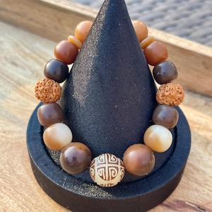 May include: A beaded bracelet with a variety of brown and tan beads, including round and textured beads. The bracelet is displayed on a black cone-shaped stand, showcasing the different colors and textures of the beads.