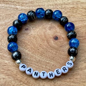May include: A beaded bracelet featuring the word "PANTHERS" spelled out in white letter beads. The bracelet also includes blue, black, and silver-colored beads. The bracelet is displayed on a wooden surface.