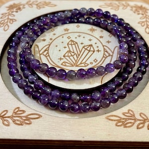 May include: Three strands of amethyst gemstone bead bracelets are displayed inside a wooden box. The beads are a deep purple color. The box has a circular cutout with a crystal design in the center.