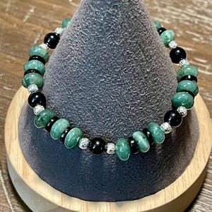 May include: A green and black beaded bracelet with silver accents. The bracelet is displayed on a gray cone-shaped stand.