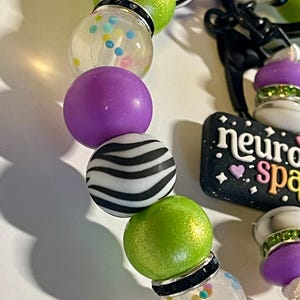 Puede incluir: Primer plano de una pulsera de cuentas con varias cuentas de colores. La pulsera presenta cuentas de color verde lima, morado, transparente y con estampado de cebra. Tambi&eacute;n es visible un dije con el texto "neuro space". Las cuentas est&aacute;n ensartadas con un cord&oacute;n negro.