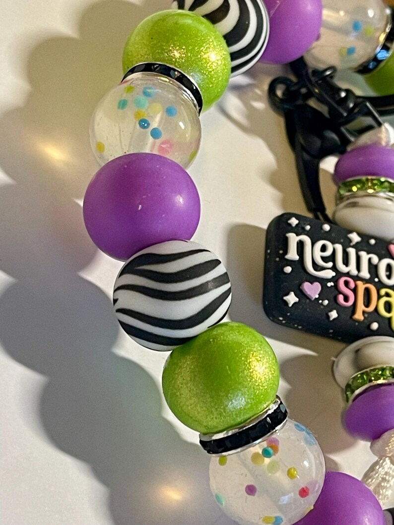 Puede incluir: Primer plano de una pulsera de cuentas con varias cuentas de colores. La pulsera presenta cuentas de color verde lima, morado, transparente y con estampado de cebra. Tambi&eacute;n es visible un dije con el texto "neuro space". Las cuentas est&aacute;n ensartadas con un cord&oacute;n negro.