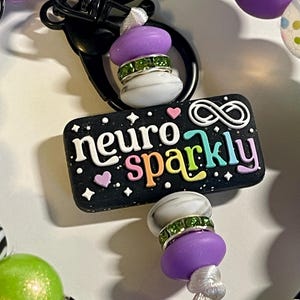 Puede incluir: Un llavero negro con las palabras "neuro sparkly" en letras coloridas. El dije est&aacute; adornado con cuentas moradas y blancas, detalles de pedrer&iacute;a verde y un cierre negro. El dije est&aacute; rodeado de otras cuentas coloridas.