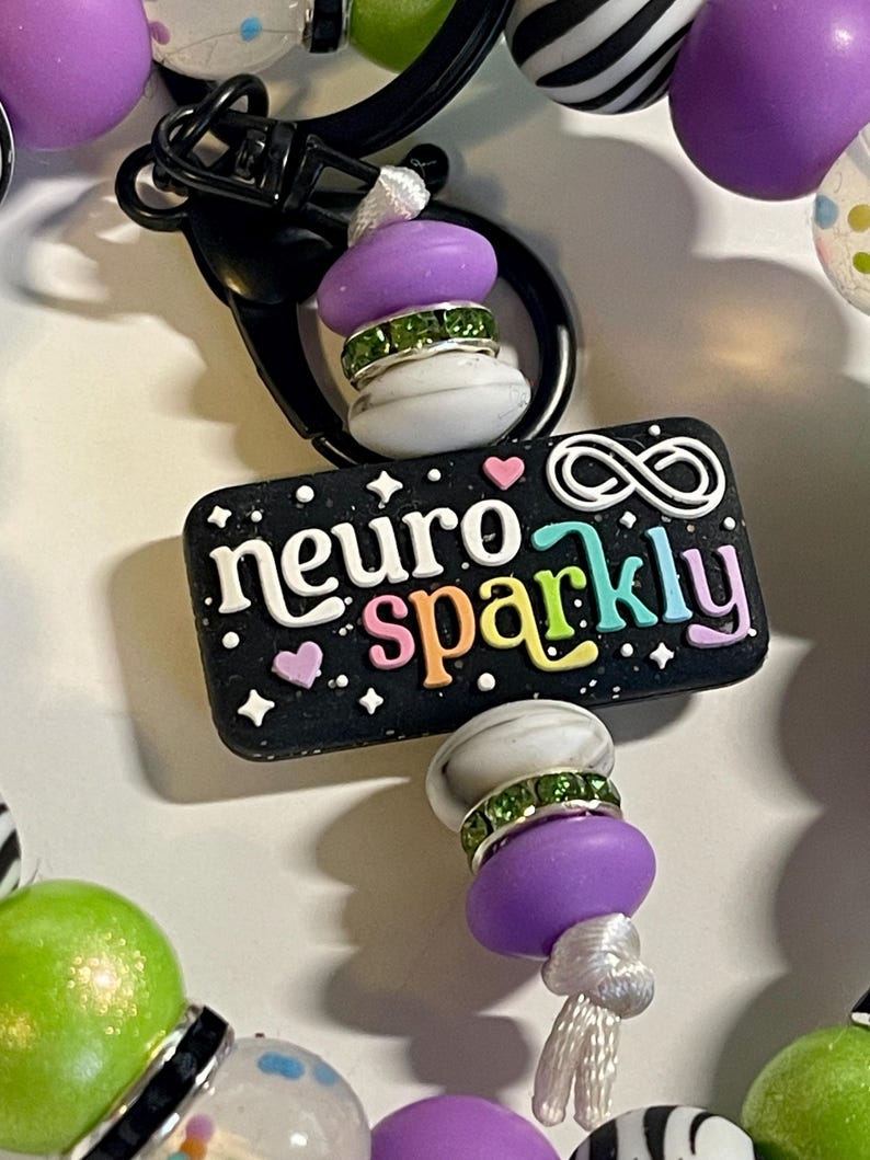 Puede incluir: Un llavero negro con las palabras "neuro sparkly" en letras coloridas. El dije est&aacute; adornado con cuentas moradas y blancas, detalles de pedrer&iacute;a verde y un cierre negro. El dije est&aacute; rodeado de otras cuentas coloridas.
