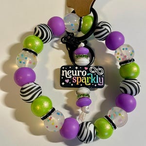 Puede incluir: Un llavero de cuentas con cuentas de colores verdes, moradas, con estampado de cebra en blanco y negro y transparentes con confeti. El llavero tiene un cierre negro y un dije que dice "neuro sparkly".