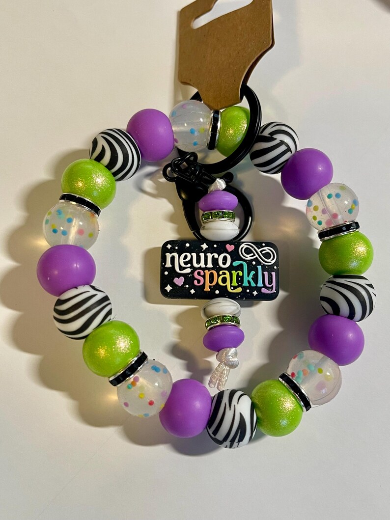 Puede incluir: Un llavero de cuentas con cuentas de colores verdes, moradas, con estampado de cebra en blanco y negro y transparentes con confeti. El llavero tiene un cierre negro y un dije que dice "neuro sparkly".