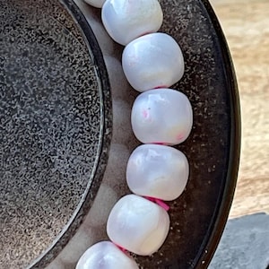 May include: A close-up of a bracelet featuring white, rounded beads with subtle pink accents. The beads are strung together with pink elastic. The bracelet is partially resting on a dark, textured surface.