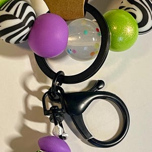 Puede incluir: Un llavero negro con un mosquet&oacute;n, con una serie de cuentas coloridas. Las cuentas incluyen esferas con estampado de cebra, moradas, transparentes con puntos de colores y verde lima. Un dije m&aacute;s peque&ntilde;o con acentos morados y verdes cuelga debajo.