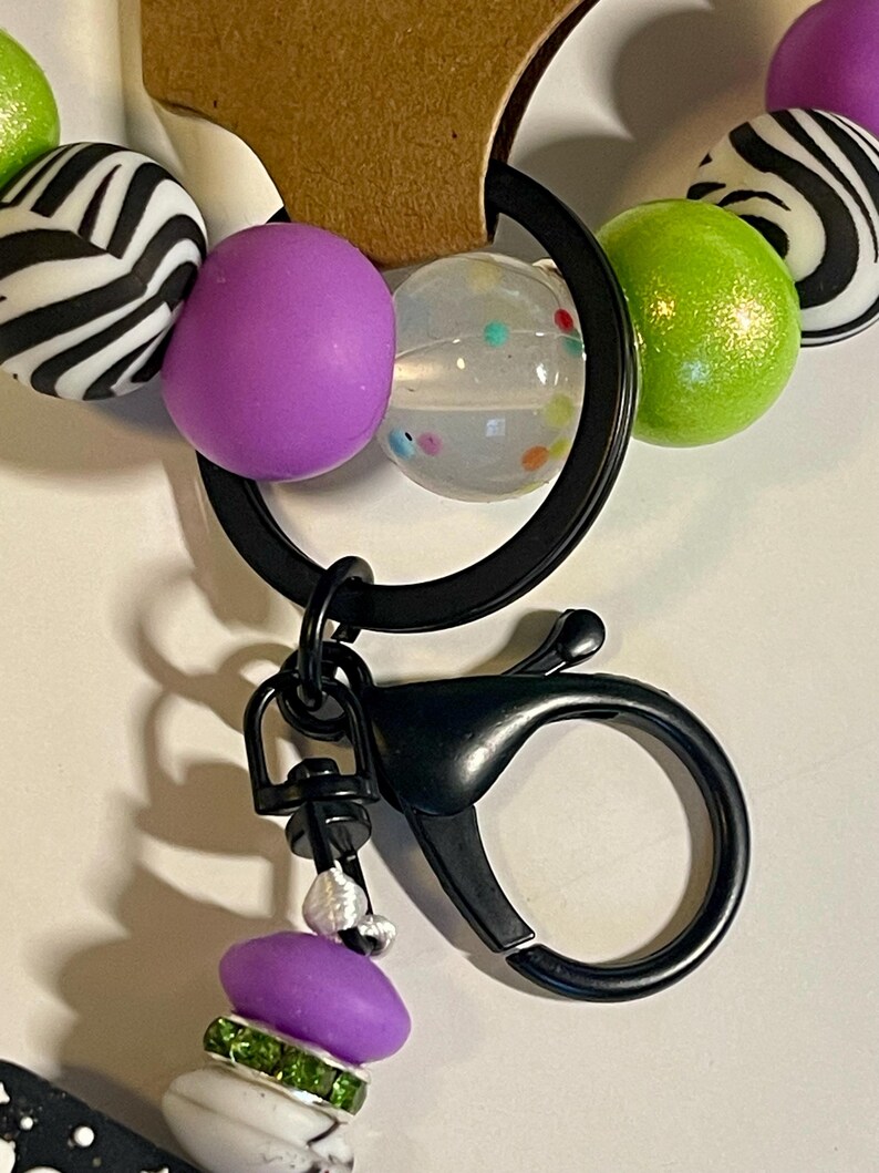 Puede incluir: Un llavero negro con un mosquet&oacute;n, con una serie de cuentas coloridas. Las cuentas incluyen esferas con estampado de cebra, moradas, transparentes con puntos de colores y verde lima. Un dije m&aacute;s peque&ntilde;o con acentos morados y verdes cuelga debajo.